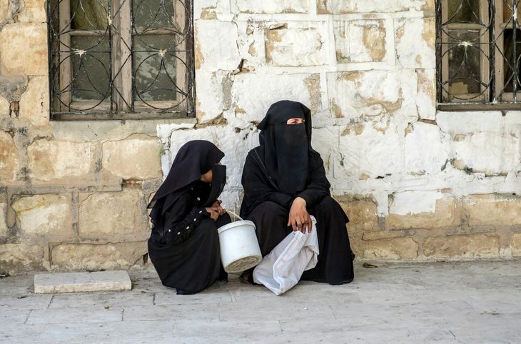 En tildækket kvinde og en pige sidder op ad en mur helt iklædt i sort, som IS-krigerne befalede. Foto: AP